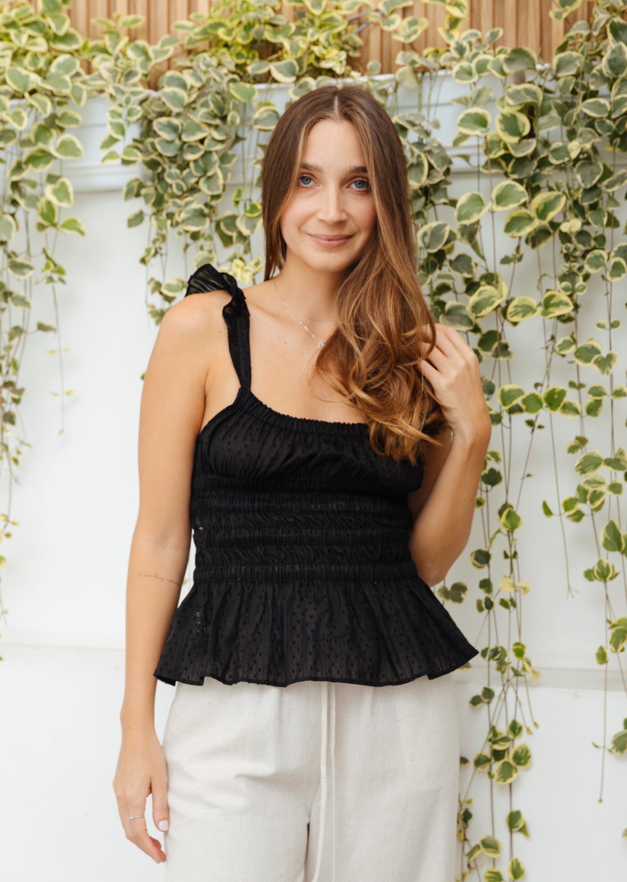 Mujer posando con top negro y pantalón blanco delante de pared con flores.